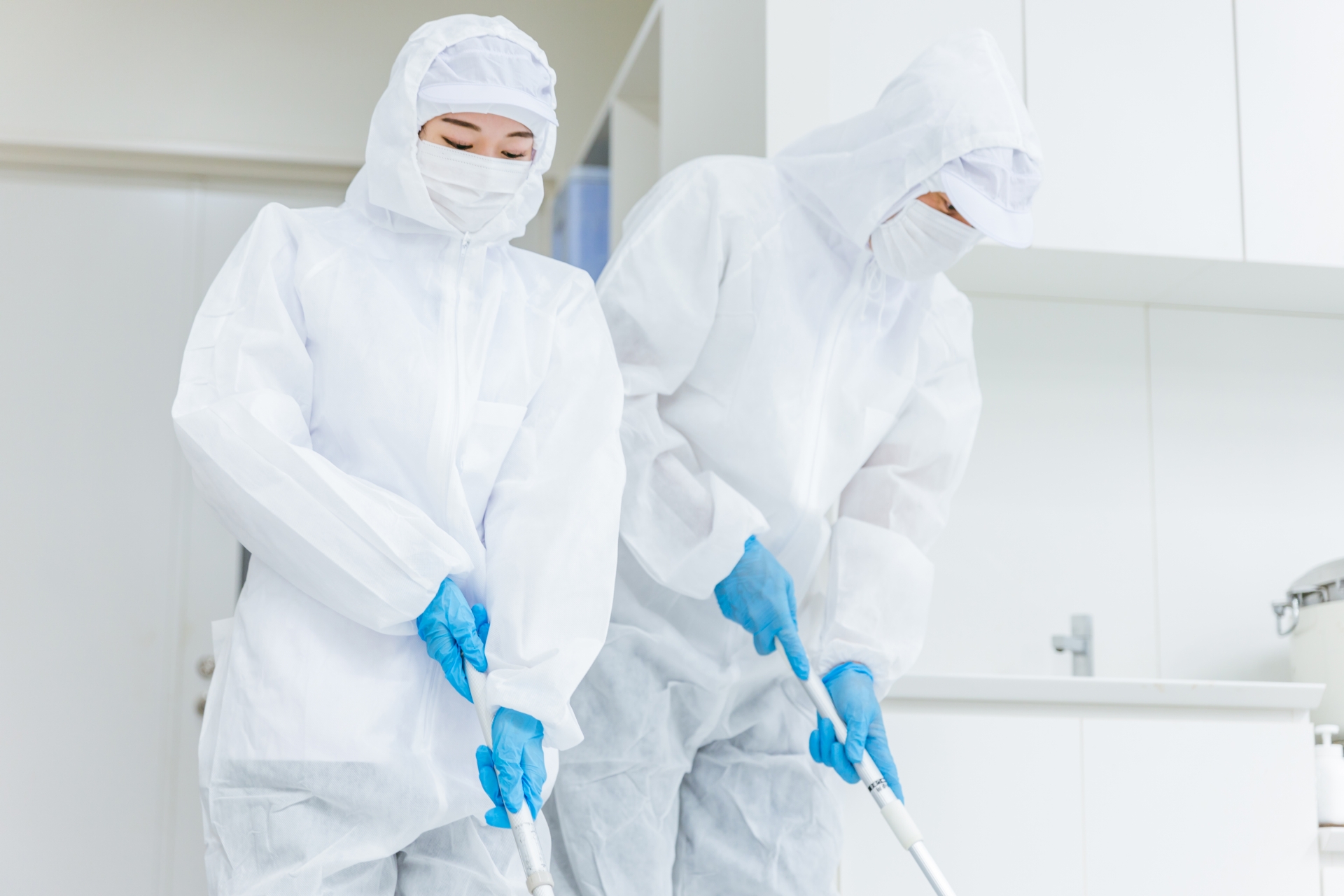Two scientists in white hazmat suits and blue gloves work at a laboratory bench, handling equipment in a cleanroom setting.