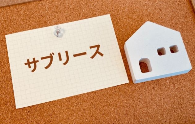 コルクボードにメモ紙に「サブリース」と書かれており、木で作られた物件が隣に置いてある写真