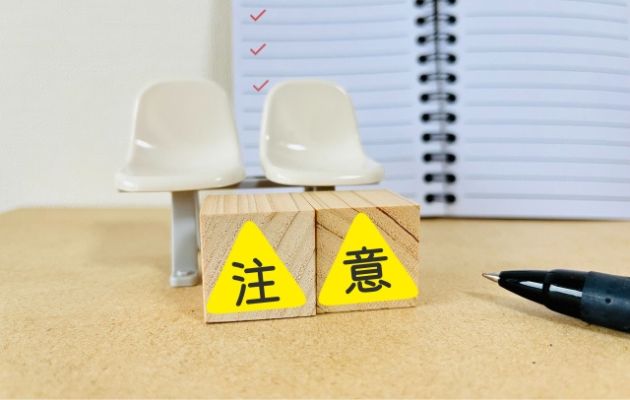 ノートにチェックマークが書かれている背景に、ミニチュアの椅子が置いてある。その下に木のブロックが置かれ、黄色い三角が書かれている。その中に「注意」と書かれている写真