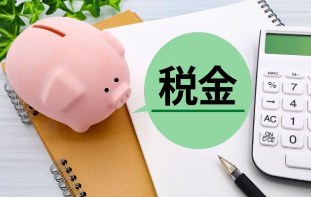 豚の貯金箱とノート、電卓、ボールペンが置いてあるところに緑の吹き出しで「税金」と書いてある写真