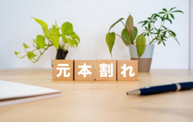 積み木に「元本割れ」と書かれている写真。背景に植物、積み木の手間にはノートとボールペンが置いてある写真