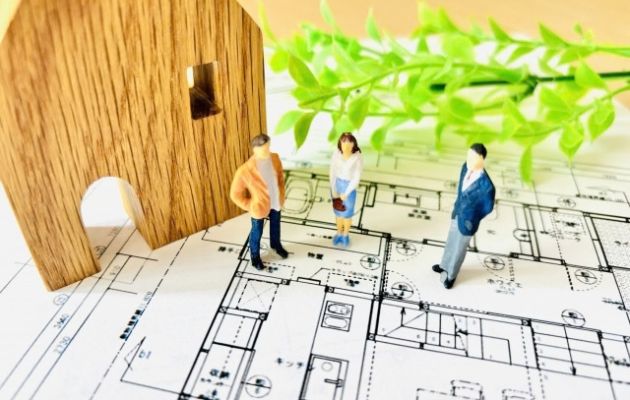 家の設計図の上に木製の家の模型と夫婦と営業マンの人形が置いてある写真
